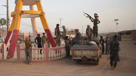 Groupe de combattants en tenue de camouflage autour d'un pick-up sur une route poussiéreuse, avec un homme en silhouette tenant un fusil automatique.