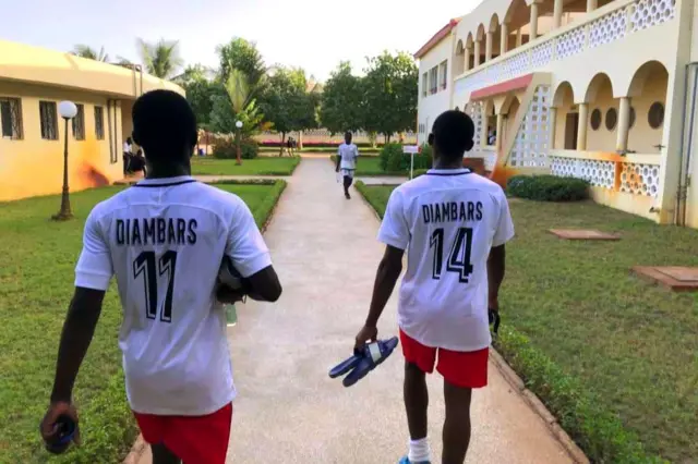 Deux joueurs en tenue blanche et short rouge marchent vers le terrain.